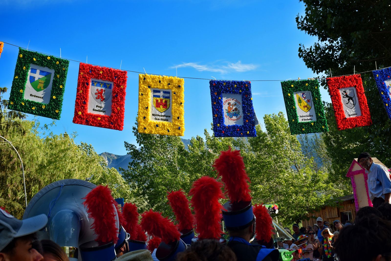 Blasons fleuris des communes suspendus au-dessus du Corso Fleuri de l’Embrunais, plumets rouges des Blackgames au premier plan