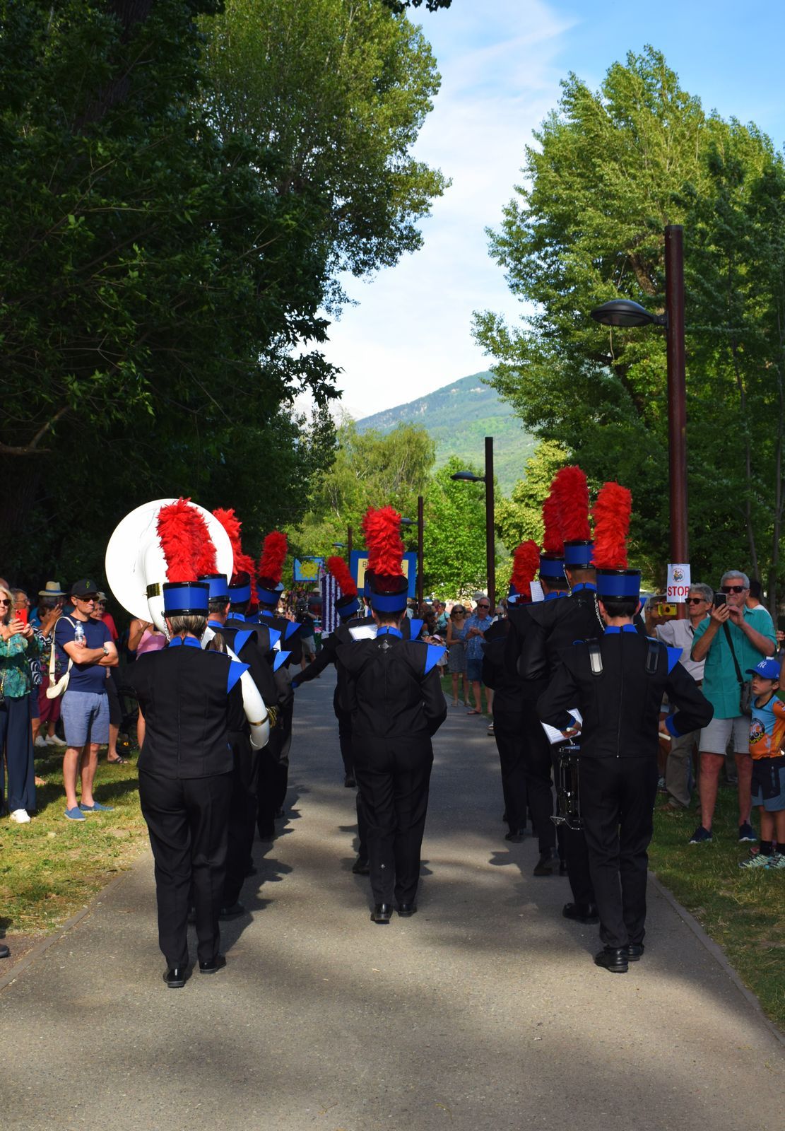 Les Marching Blackgames défilent en formation sur le parcours du corso, shakos à plumets rouges, foule et montagne en arrière-plan